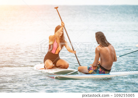 Sea woman and man on sup. Silhouette of happy young woman and man, surfing on SUP board, confident paddling through water surface. Idyllic sunset. Active lifestyle at sea or river. Sea woman and man on sup. Silhouette of happy young woman and man, surfing on SUP board, confident paddling through water surface. Idyllic sunset. Active lifestyle at sea or river. 109255005
