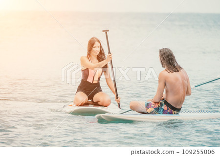 Sea woman and man on sup. Silhouette of happy young woman and man, surfing on SUP board, confident paddling through water surface. Idyllic sunset. Active lifestyle at sea or river. Sea woman and man on sup. Silhouette of happy young woman and man, surfing on SUP board, confident paddling through water surface. Idyllic sunset. Active lifestyle at sea or river. 109255006