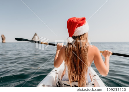 Woman in kayak back view. Happy young woman in Santa hat floating in kayak on calm sea. Summer holiday vacation and cheerful female people relaxing having fun on the boat. 109255320