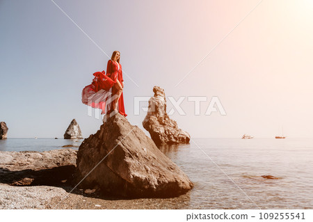 Woman travel sea. Young Happy woman in a long red dress posing on a beach near the sea on background of volcanic rocks, like in Iceland, sharing travel adventure journey Woman travel sea. Young Happy woman in a long red dress posing on a beach near the sea on background of volcanic rocks, like in Iceland, sharing travel adventure journey 109255541