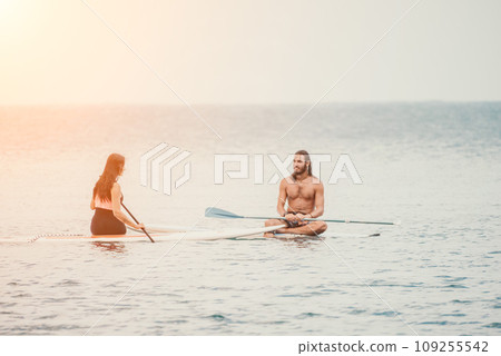 Sea woman and man on sup. Silhouette of happy young woman and man, surfing on SUP board, confident paddling through water surface. Idyllic sunset. Active lifestyle at sea or river. 109255542
