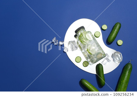 A bottle filled with cucumber detox water placed on a mirror, decorated with round slices of cucumber and ices. Blank label bottle for product mockup advertising A bottle filled with cucumber detox water placed on a mirror, decorated with round slices of cucumber and ices. Blank label bottle for product mockup advertising 109256234