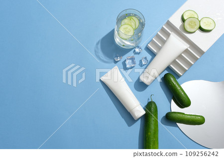 Two white tubes decorated with a staircase, ices and a mirror. A glass of cucumber detox water is featured. Skin care concept with empty label for Cucumber extracted product Two white tubes decorated with a staircase, ices and a mirror. A glass of cucumber detox water is featured. Skin care concept with empty label for Cucumber extracted product 109256242