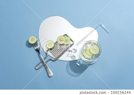 A mirror arranged with cucumber slices, grater, straw, fork and a glass of water. Cucumber (Cucumis sativus) is a food that contains many vitamins and minerals 109256244