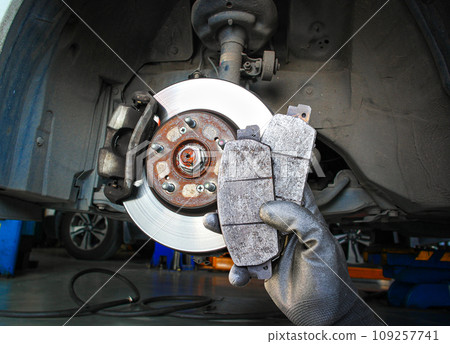 Used car brake pads in the hands of a mechanic with brake discs and brake calipers on the background Used car brake pads in the hands of a mechanic with brake discs and brake calipers on the background 109257741