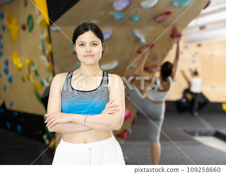 Positive slender Asian girl in sportswear is standing near climbing wall in gym 109258660
