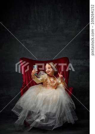 Little princess, girl sitting on royal throne and taking selfie showing piece sign against dark vintage background. 109264152