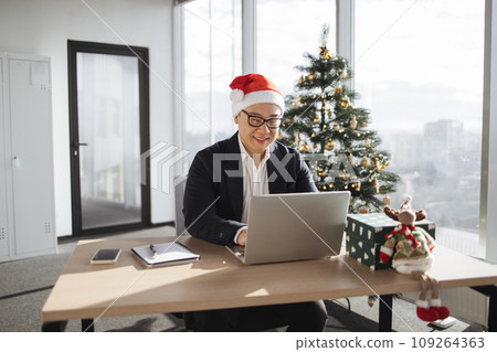 Businessman sitting in office near xmas tree working on laptop Businessman sitting in office near xmas tree working on laptop 109264363