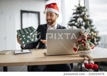 Focus on Christmas deer on table, blurred background of smiling Caucasian man in Santa hat. 109264476