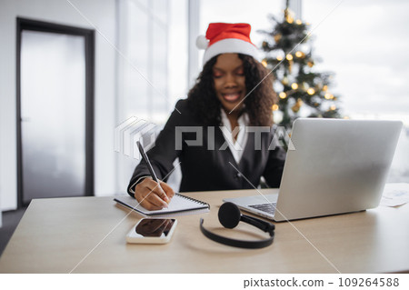 African businesswoman in a Santa hat with pen making notes. 109264588