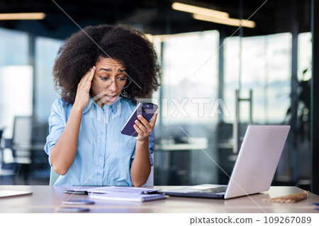 Upset frustrated and sad businesswoman, got bad news online on phone, female employee reading news nervously, working inside office with laptop, woman with curly hair depressed. 109267089