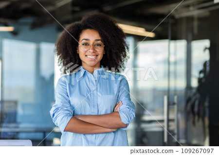 Businesswoman with arms crossed inside office at workplace, woman smiling and looking at camera, female worker with curly hair and glasses near window. Businesswoman with arms crossed inside office at workplace, woman smiling and looking at camera, female worker with curly hair and glasses near window. 109267097