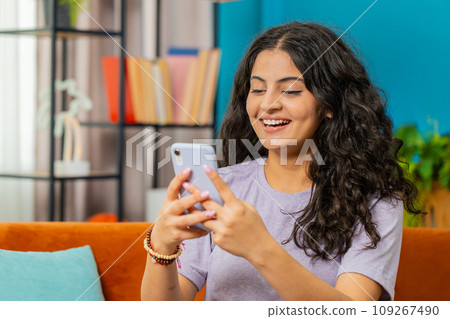 Indian Arabian young woman sitting on sofa use smartphone share messages on social media application Indian Arabian young woman sitting on sofa use smartphone share messages on social media application 109267490
