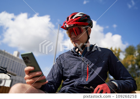 Man in a bicycle helmet sits on a bench and looks at his phone, resting while cycling in the city Man in a bicycle helmet sits on a bench and looks at his phone, resting while cycling in the city 109268258