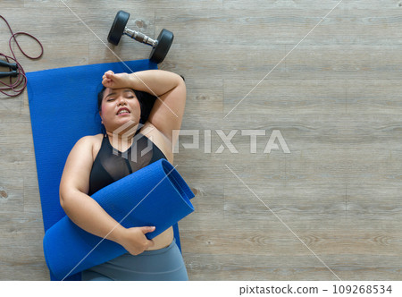 A plus-size woman lies exhausted on a blue mat, taking a breather after a vigorous workout session with dumbbell and jump rope. Top view 109268534