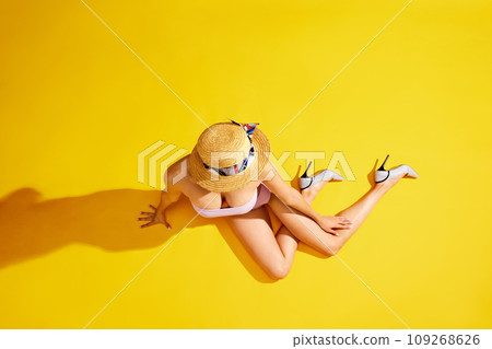 Slim young woman, in swimsuit, straw hat and heels lying against yellow studio background. Top view 109268626