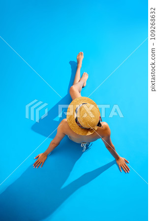 Top view. Elegant young woman with slim, fit, tanned body, wearing swimsuit and straw hat, lying against blue studio background 109268632