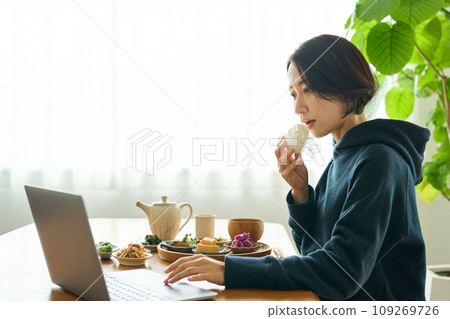 A woman hurriedly eats breakfast while looking at a computer A woman hurriedly eats breakfast while looking at a computer 109269726