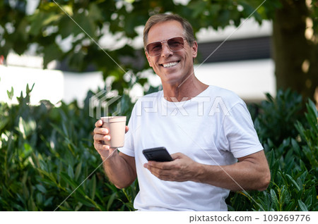 Middle aged Caucasian man using smartphone drinking coffee outdoors in city park. 109269776