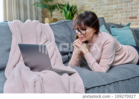 Happy middle aged woman lying at home on sofa in living room using laptop Happy middle aged woman lying at home on sofa in living room using laptop 109270027