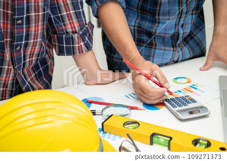 Asian engineer and partner team learn and talk with house plan project blueprint, graph and yellow helmet and engineering construction tools on table work. Asian engineer and partner team learn and talk with house plan project blueprint, graph and yellow helmet and engineering construction tools on table work. 109271373
