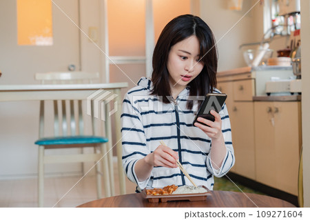 A young woman eating a convenience store lunch at home while using a smartphone A young woman eating a convenience store lunch at home while using a smartphone 109271604