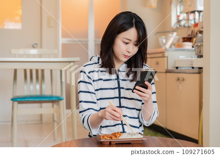 A young woman eating a convenience store lunch at home while using a smartphone A young woman eating a convenience store lunch at home while using a smartphone 109271605