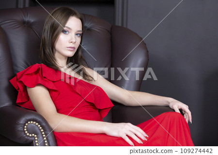 Beautiful model in an armchair. Luxury girl is sitting on a leather chair. Young woman in a red dress 109274442