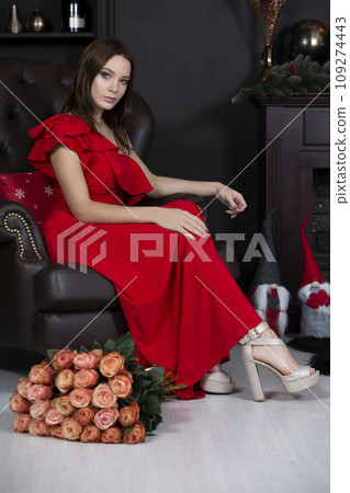 Vertical beautiful model in an armchair. Luxury girl sitting on a leather chair with a bouquet of roses. Young woman in a red dress 109274443