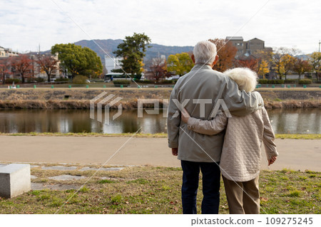 A friendly elderly couple standing shoulder to shoulder on the riverbank 109275245