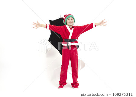 Child Santa Claus comes out of a hole in paper. A boy in a New Year's costume is smiling. Holiday surprise. Christmas holiday concept Child Santa Claus comes out of a hole in paper. A boy in a New Year's costume is smiling. Holiday surprise. Christmas holiday concept 109275311