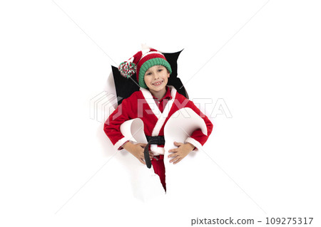Child Santa Claus looks out of a hole in paper. A boy in a New Year's costume is smiling. Holiday surprise. Christmas holiday concept 109275317