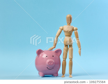 Ceramic pink piggy bank and a wooden puppet doll on a blue background. Concept of increasing income from bank accounts 109275564