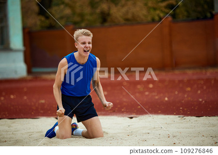 Professional sportsman screams rejoicing at successful completion of long jump and victory while kneeling in squeak. Professional sportsman screams rejoicing at successful completion of long jump and victory while kneeling in squeak. 109276846