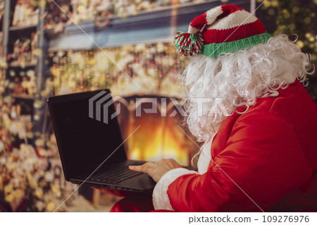 Santa Claus is looking at the laptop. A man in a Christmas suit at the computer on the background of New Year's lights. The modern wizard. Christmas holidays concept 109276976