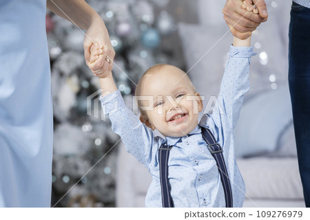 Happy baby with parents at christmas. Hands of mom and dad hold the child against the background of the Christmas tree. The face of a little boy in the New Year. Family in the winter holidays. 109276979