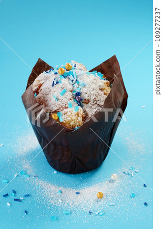 Cupcake with icing sugar and sprinkles on a blue background, vertical photo, close-up. Backdrop with a decorated cupcake wrapped in brown butter paper, vertical picture. 109277237