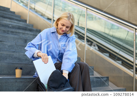 Portrait of young modern woman, student freelancer, sitting on public stairs outdoors, putting away her laptop, packing backpack, drinking coffee outside Portrait of young modern woman, student freelancer, sitting on public stairs outdoors, putting away her laptop, packing backpack, drinking coffee outside 109278014