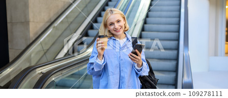 Image of beautiful female model, student standing near escalator, raising cup of takeaway coffee, smiling and looking happy, has mobile phone in hand and backpack on shoulder 109278111