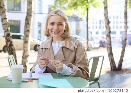 Image of young blonde woman in stylish trench, sitting in outdoor cafe, drinking coffee and working on documents, studying, doing homework in her notebook, smiling at camera Image of young blonde woman in stylish trench, sitting in outdoor cafe, drinking coffee and working on documents, studying, doing homework in her notebook, smiling at camera 109278140