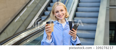 Portrait of young beautiful woman, girl student with cup of coffee and smartphone, standing near escalator in city centre, smiling and looking at camera 109278217