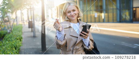 Lovely young female model in trench coat, showing heart gesture with fingers, holding smartphone and smiling at camera, standing on empty sunny street 109278218
