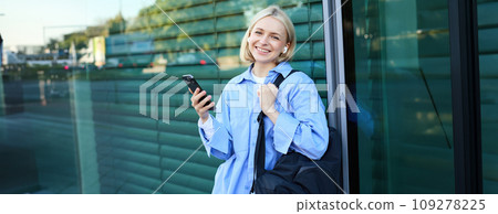 Lifestyle portrait of smiling young female model, student with backpack, waiting for someone on street, standing outdoors with smartphone 109278225