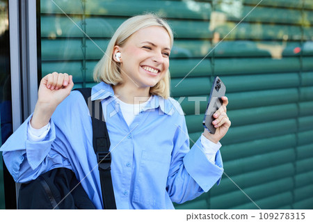 Carefree woman with backpack and mobile phone, wearing wireless earphones, listening to music and smiling, standing on street in city centre, laughing 109278315
