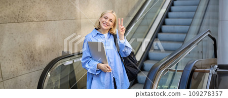 Smiling, beautiful girl student, woman with laptop and backpack, using escalator in city, showing peace, v-sign gesture, looking happy 109278357