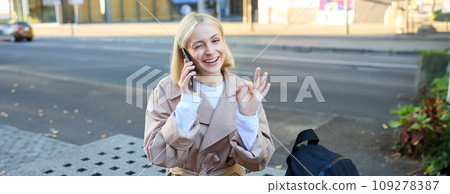 Image of young woman talking on mobile phone, sitting on bench outside, showing okay, ok approval gesture, made a deal over the telephone 109278387