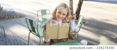 Portrait of young beautiful blond woman, artsy girl in coffee shop, holding notebook and pen, writing in her journal, drawing sketches outdoors 109278390