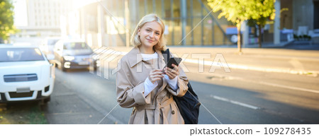 Lifestyle portrait of young woman on street, standing near road with cars, carries backpack and holds mobile phone, requests a ride on smartphone app, smiling and looking happy at camera Lifestyle portrait of young woman on street, standing near road with cars, carries backpack and holds mobile phone, requests a ride on smartphone app, smiling and looking happy at camera 109278435