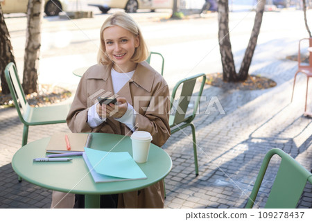 Image of young beautiful girl, student studying, doing homework in cafe, using mobile phone, holding smartphone, smiling 109278437