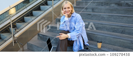 Portrait of young modern woman, student or freelancer, sitting on outdoor stairs, resting in city, has laptop and coffee, looking at camera with confident, smiling face Portrait of young modern woman, student or freelancer, sitting on outdoor stairs, resting in city, has laptop and coffee, looking at camera with confident, smiling face 109278438